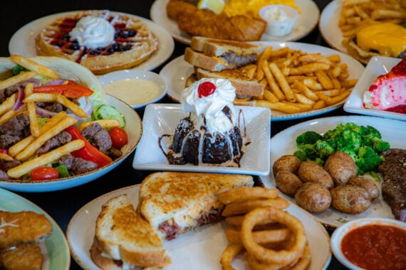 A variety of dishes on a table, including burgers, fries, grilled cheese, onion rings, salad, broccoli, potatoes, fried fish, waffles with berries, and a chocolate dessert topped with whipped cream and a cherry.