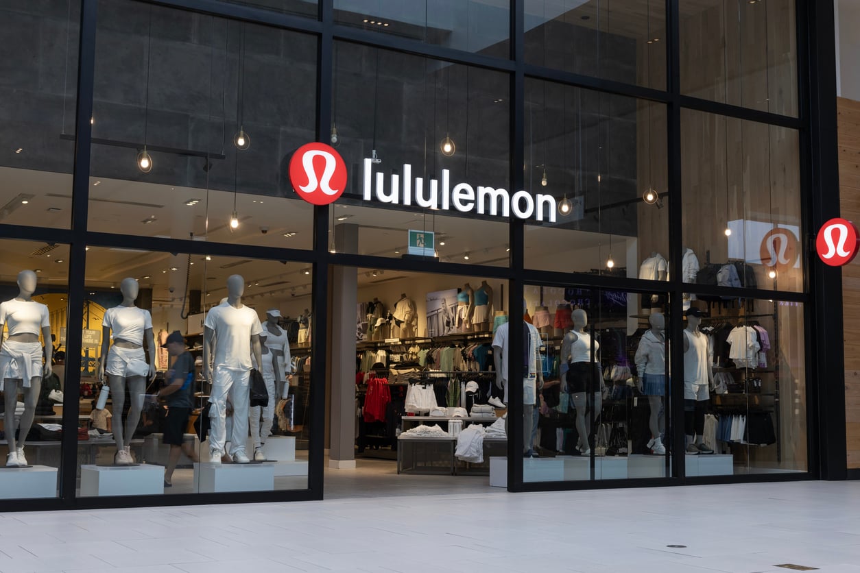 A Lululemon store with large glass windows displays mannequins dressed in athletic wear. The illuminated Lululemon logo and store name are above the entrance, and shelves of clothing are visible inside.
