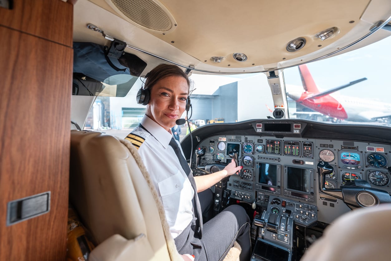 A female pilot wearing a uniform and headset sits in the cockpit of an airplane, looking back and smiling at the camera, with aircraft controls and another plane visible outside the window.