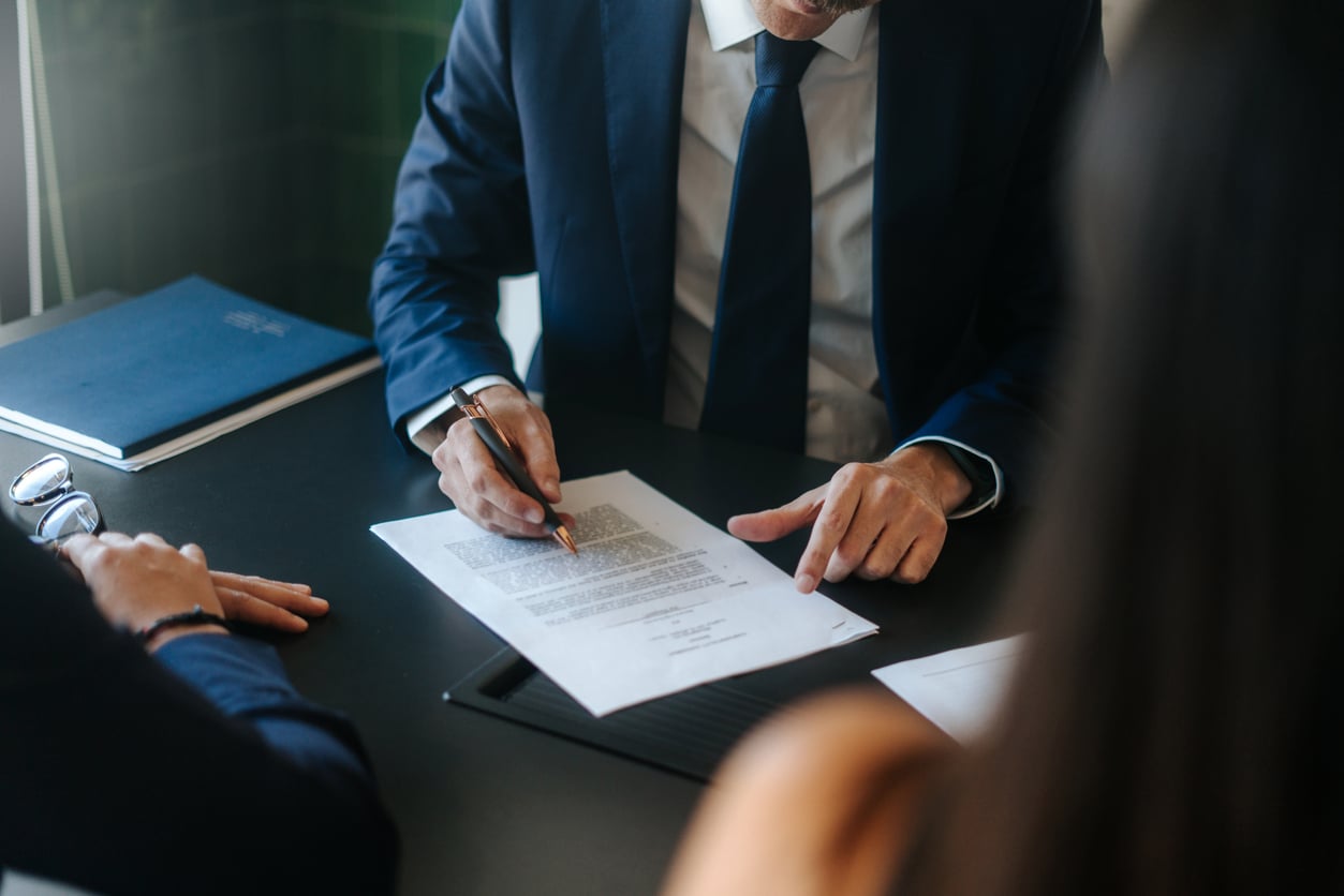 A person in a suit points to a section of a printed document on a table, discussing it with two other people whose faces are not visible. A closed blue folder lies nearby.