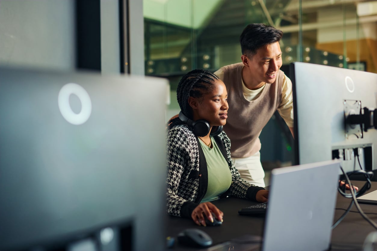 Two people working together at a computer desk; one is sitting and using a keyboard and mouse while the other stands beside, smiling and looking at the monitor. Computer screens and headphones are visible.
