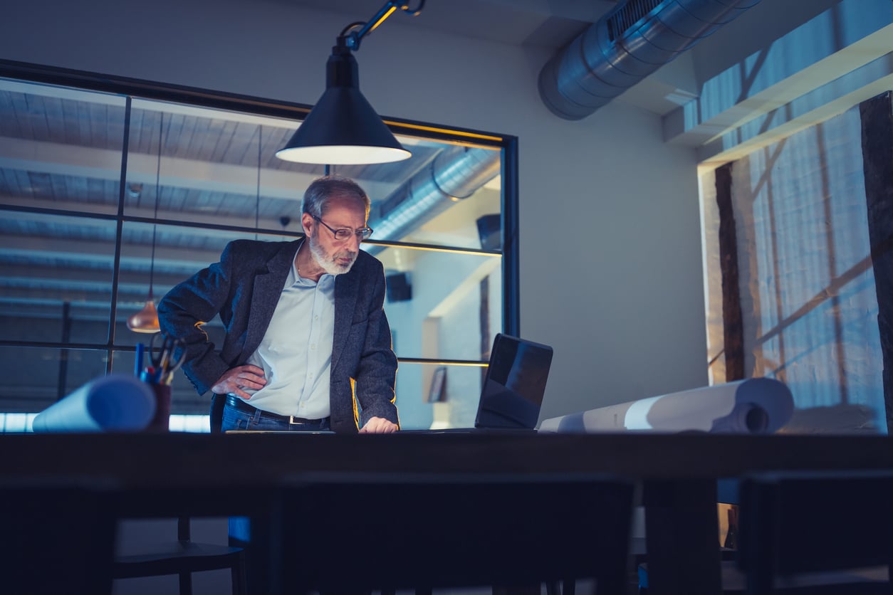 An older man in a blazer leans over a table with architectural blueprints, looking at a laptop in a modern office with large windows and industrial lighting.