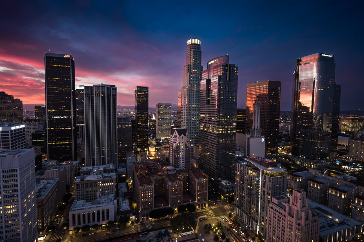 Downtown cityscape at dusk with tall skyscrapers lit by office lights, under a vibrant sky with deep blue and pink clouds. Streets and buildings are illuminated as night falls.