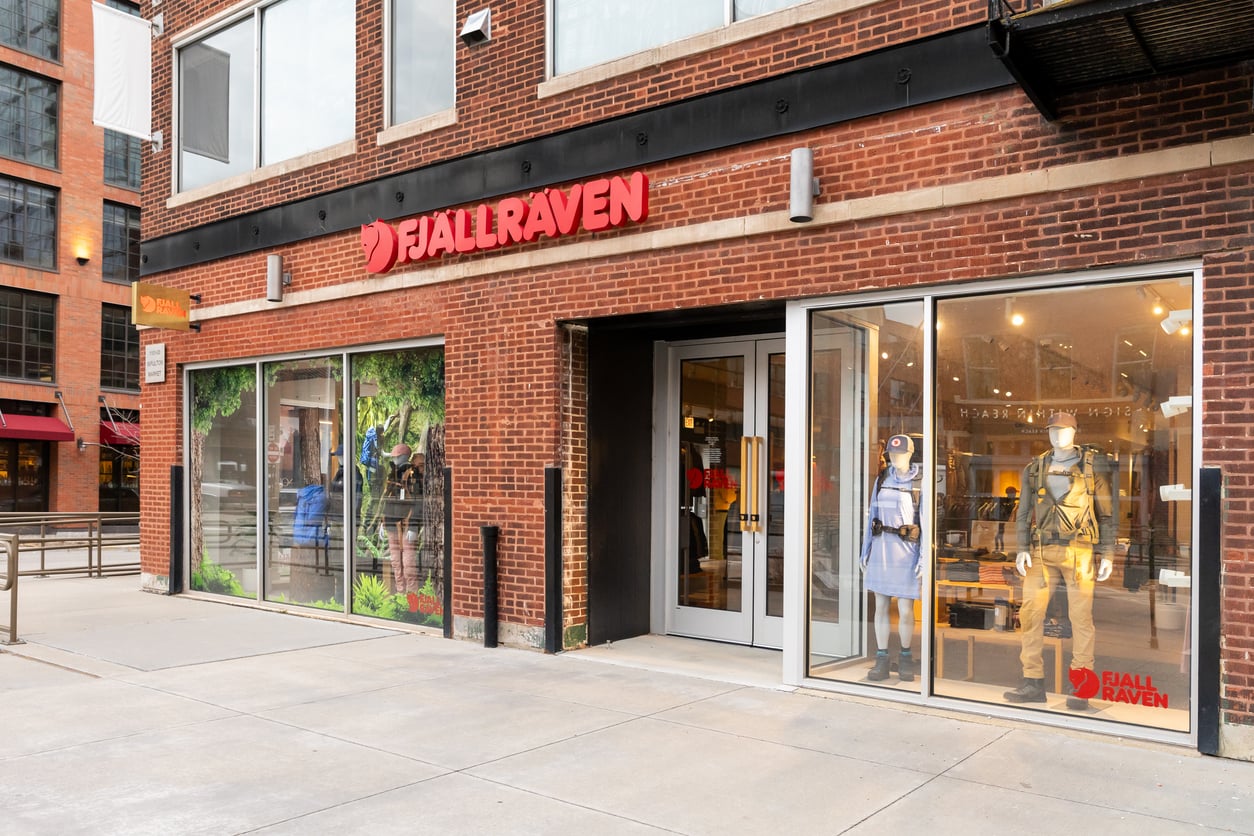 The exterior of a Fjällräven store with large glass windows displaying mannequins in outdoor apparel. The red Fjällräven sign is above the entrance, and the storefront is set in a brick building on a city sidewalk.