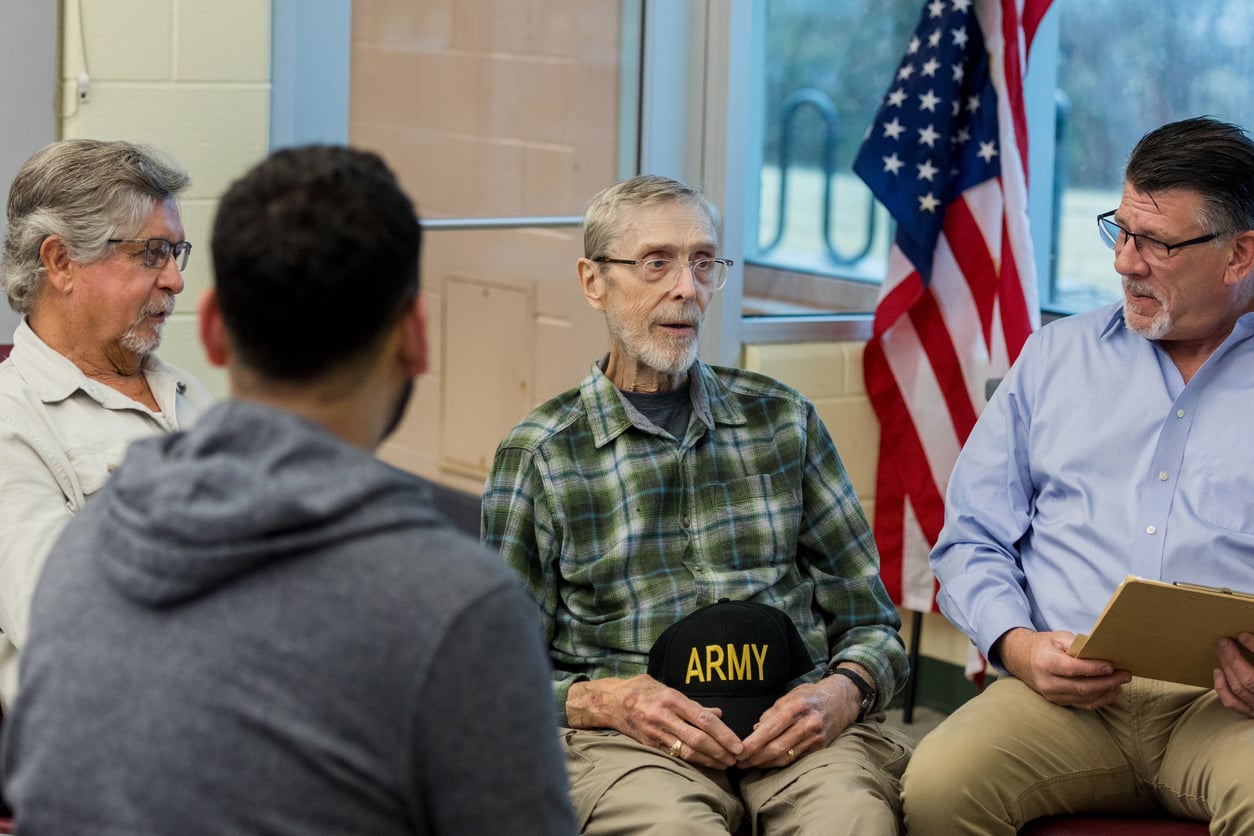 Four men sit in a discussion circle indoors; one older man in a plaid shirt holds a cap with "ARMY" on it. An American flag is visible in the background near large windows.