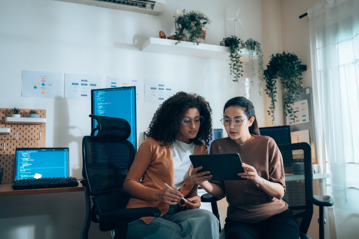Two women sit together in an office, looking at a tablet. Computer monitors with code are on desks nearby. The room has plants, shelves, and large windows letting in natural light.
