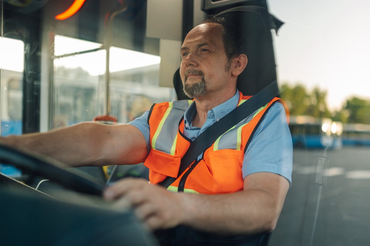 A middle-aged man wearing a reflective orange safety vest and blue shirt drives a vehicle, likely a bus, while looking out the windshield in daylight.