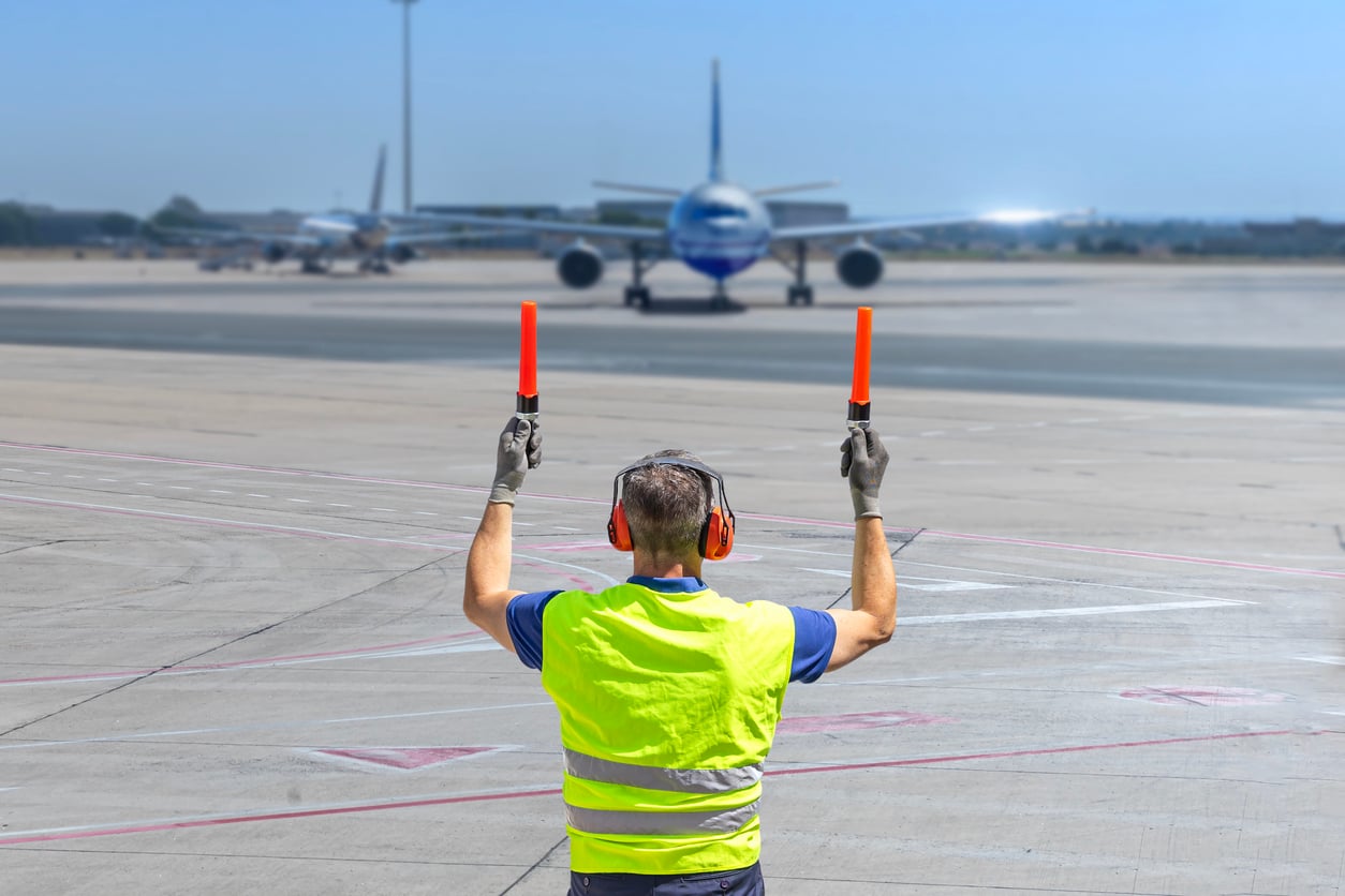 A ground crew worker in a reflective vest and earmuffs directs an approaching airplane on the airport tarmac using two orange signal batons.