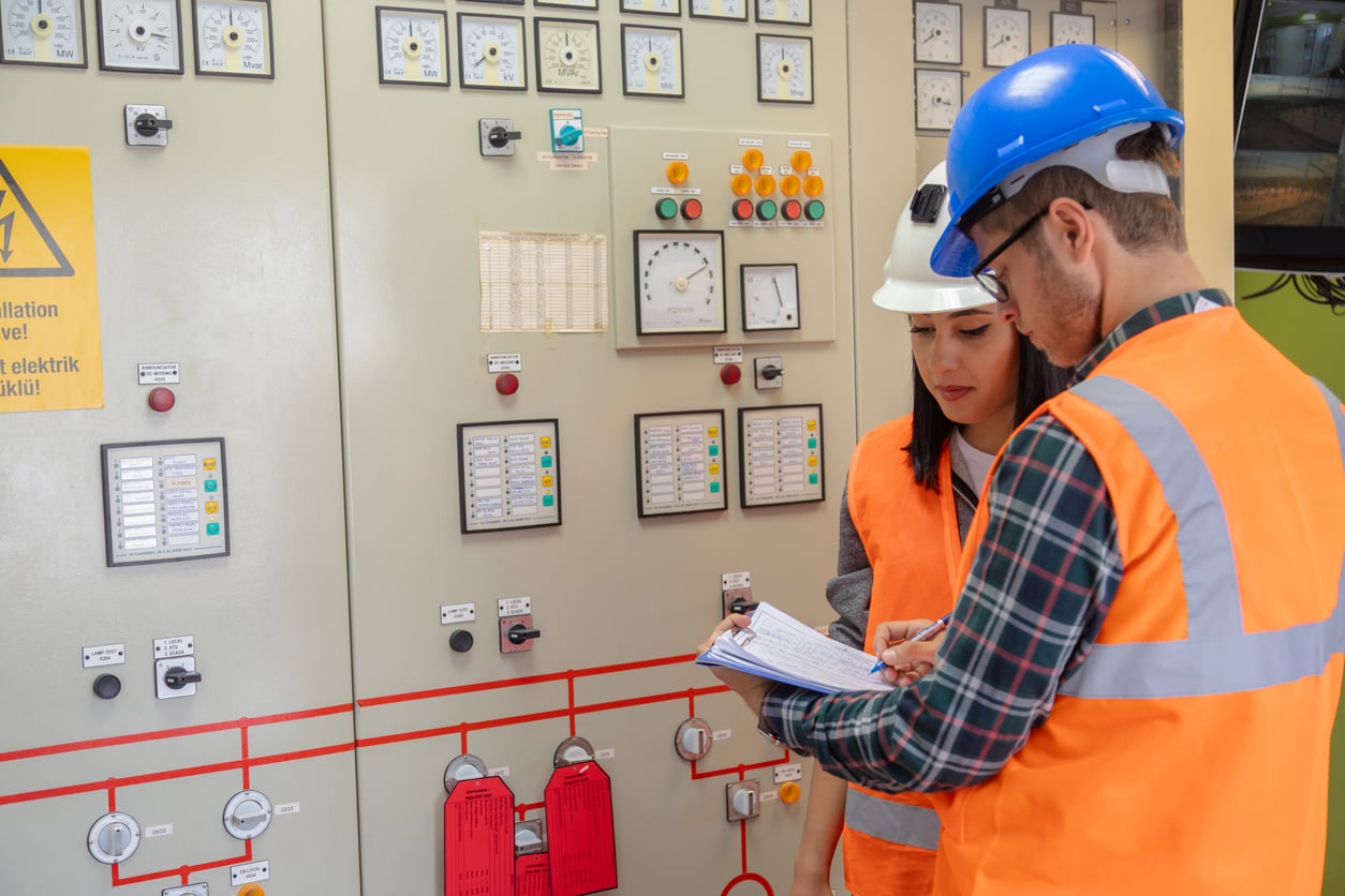 Two engineers wearing hard hats and orange safety vests review notes in front of a control panel with dials, gauges, and warning signs in an industrial setting.