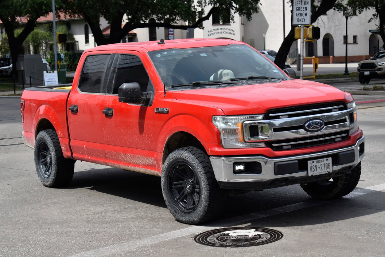 A red Ford F-150 pickup truck with black wheels and some mud on its sides is parked on a city street. Trees and buildings are visible in the background.