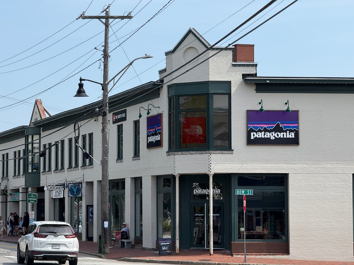 A corner brick building with large windows houses a Patagonia store. The store's sign with the mountain logo is visible above the entrance. A white SUV is parked along the street and a few pedestrians walk nearby.