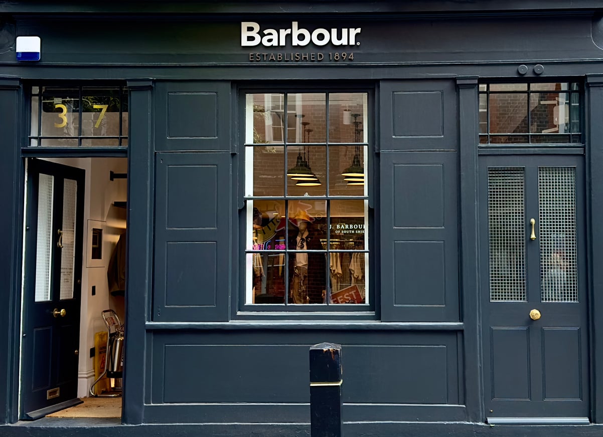 Exterior of a dark blue Barbour store with gold address number 37, large front windows, and a glass door. The Barbour logo and "Established 1894" are displayed above the entrance.
