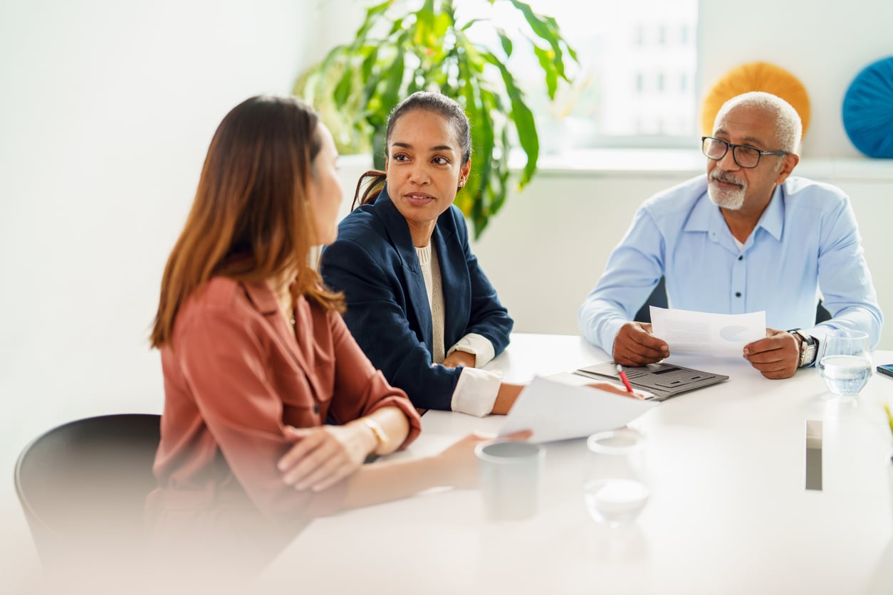 Three people sit at a table in a bright office, engaged in discussion. One woman faces the camera while talking to another woman; an older man with papers listens attentively. A plant and window are in the background.