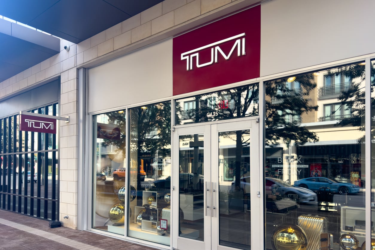 Storefront of a TUMI retail shop with large glass windows and doors, displaying luggage inside. The TUMI logo is visible above the entrance and on a sign to the left. Reflections of trees and buildings can be seen in the glass.