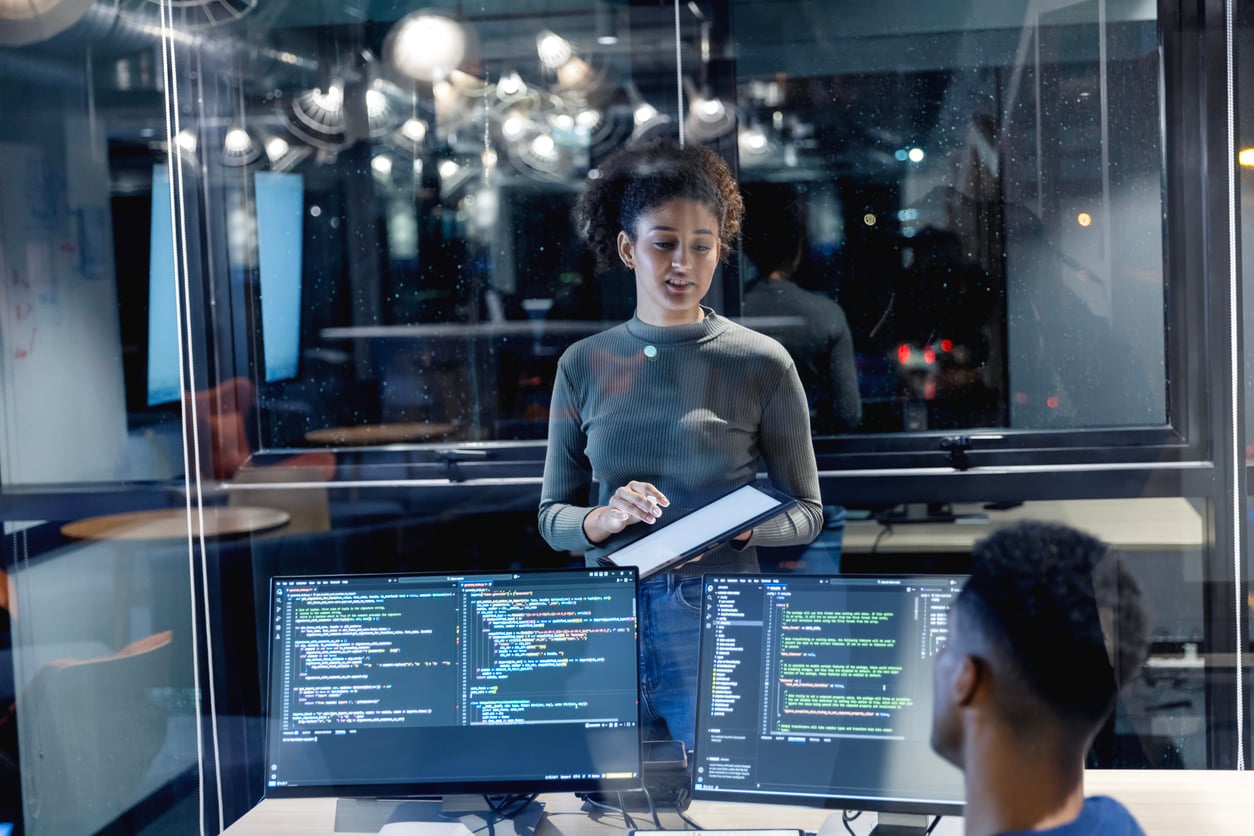 A woman holding a tablet stands in front of a window, talking to a seated man. Two computer monitors display code on their screens in the foreground, indicating a collaborative tech or programming environment.