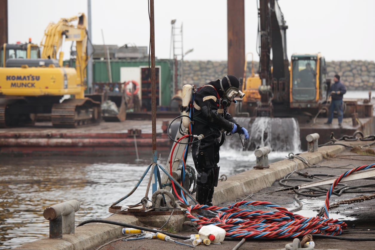 A diver in full gear stands on a dock next to colorful coiled hoses and industrial equipment, preparing for underwater work at a construction site near the water. Excavators and machinery are visible in the background.