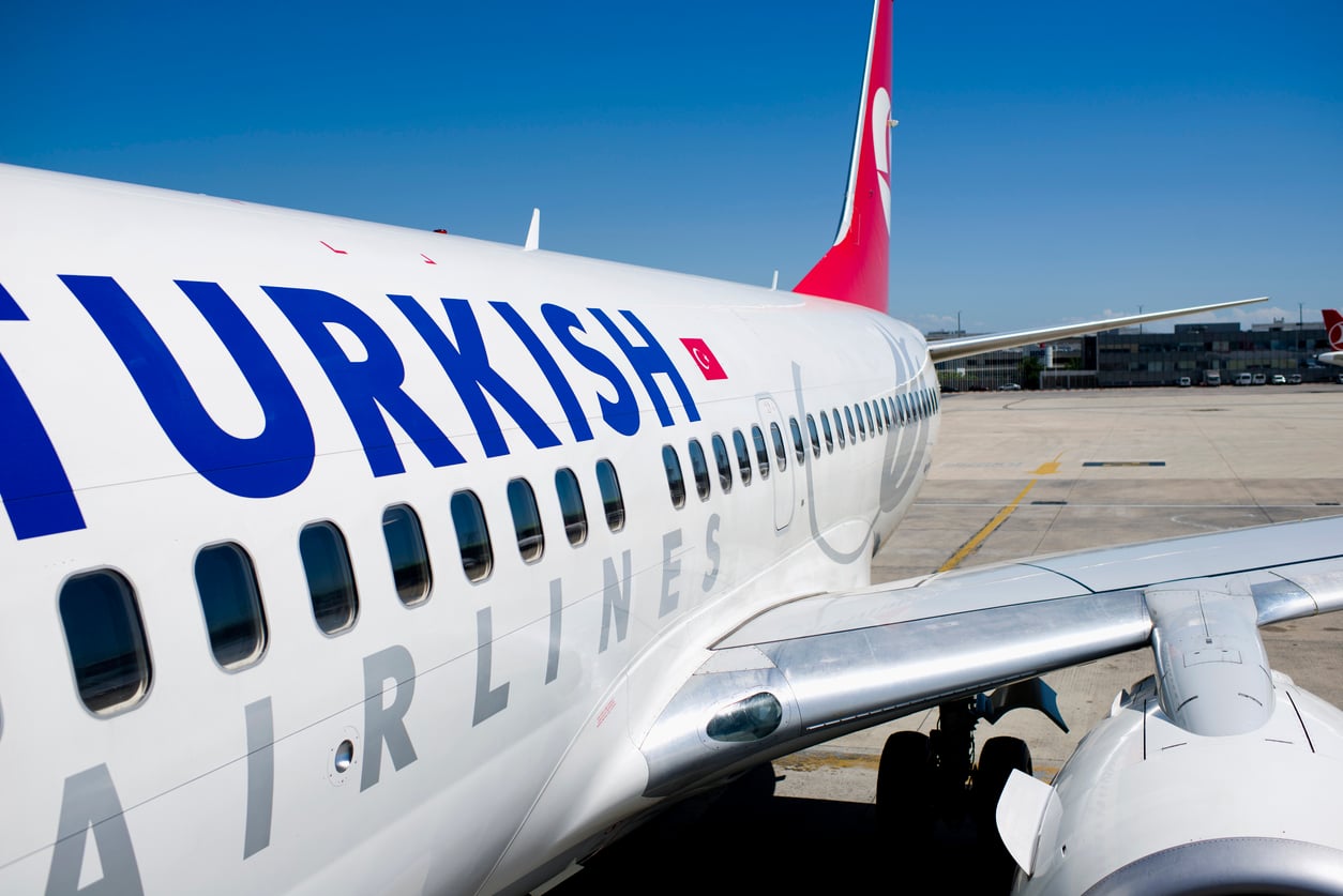 Close-up view of a Turkish Airlines airplane on the tarmac, showing the company logo on the fuselage and part of the red tail fin against a clear blue sky.