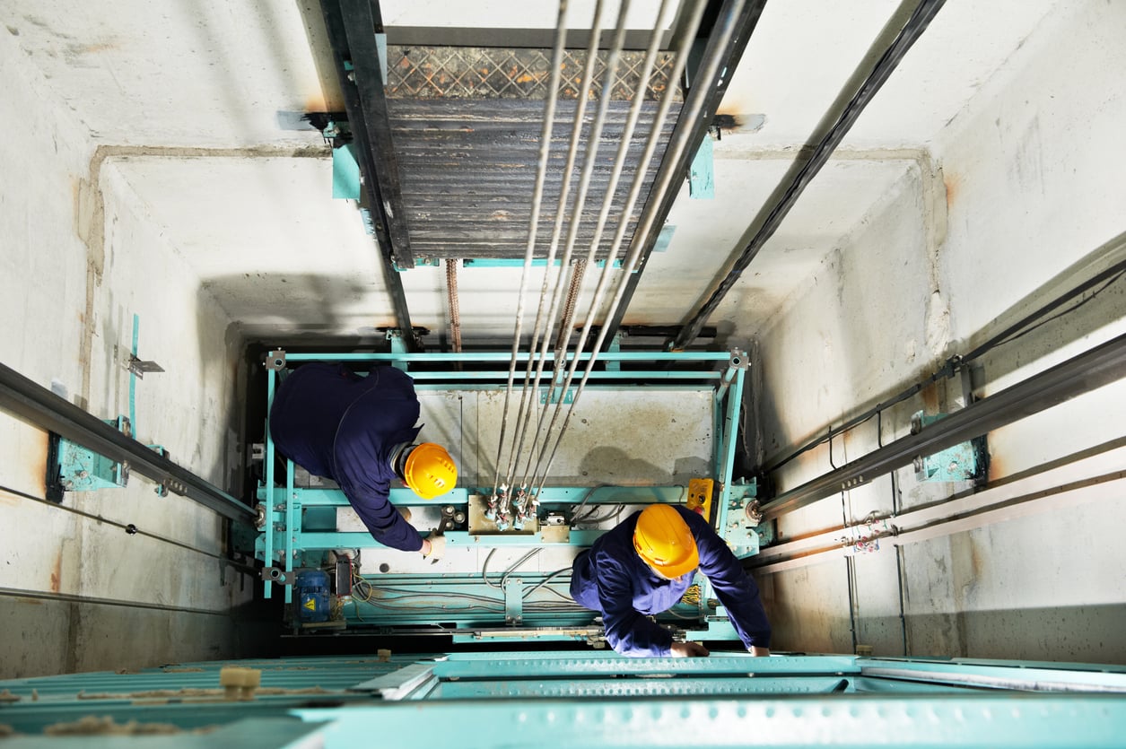 Two workers wearing yellow helmets and blue uniforms are repairing or inspecting machinery inside an elevator shaft, viewed from above. Cables and elevator components are visible around them.