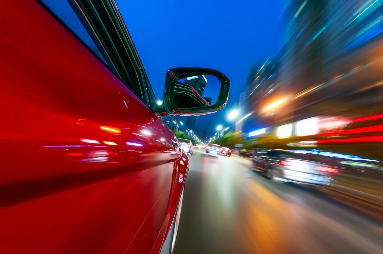 View from the side of a red car driving fast through a city at night, with blurred lights and streaks showing motion, and other cars visible ahead on the brightly lit road.