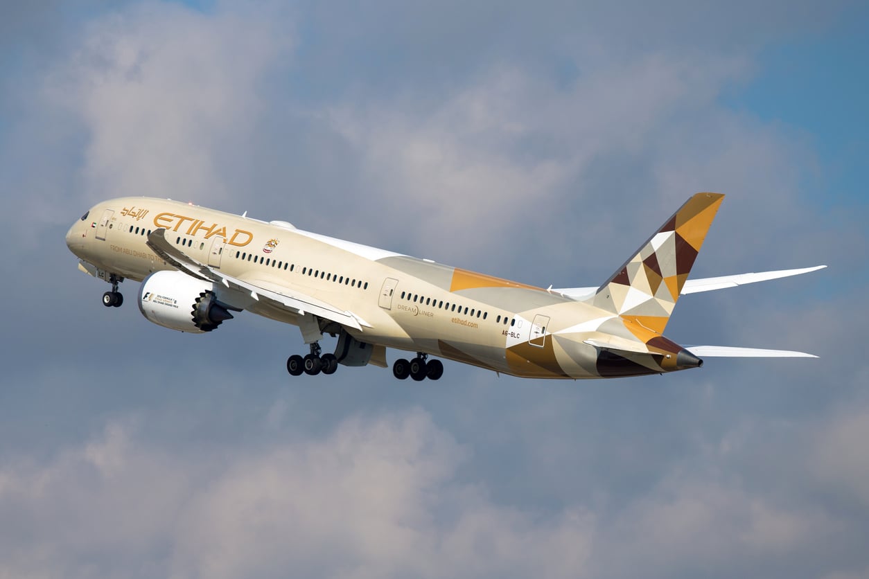 Etihad Airways Boeing 787 Dreamliner aircraft taking off into a partly cloudy sky, showing its gold and beige geometric livery.