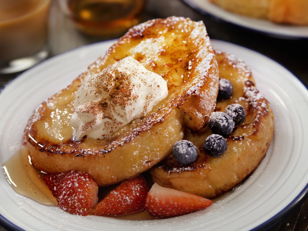Two slices of French toast topped with whipped cream, powdered sugar, and cinnamon, served with fresh strawberries and blueberries, and drizzled with syrup on a white plate.