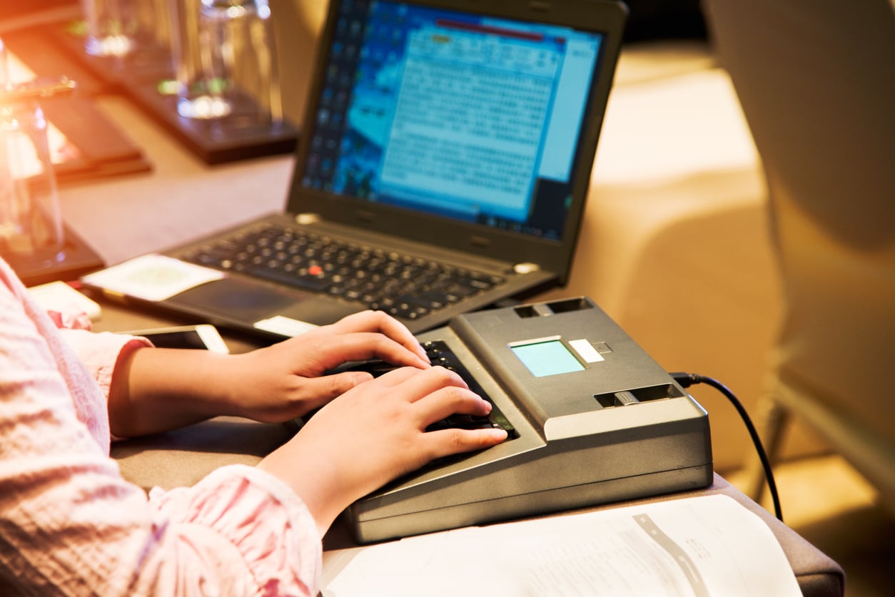 A person types on a stenotype machine with both hands while a laptop with a document open sits in the background on a desk.
