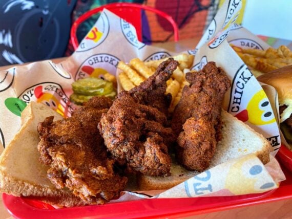 A red tray with spicy fried chicken on white bread, crinkle-cut fries, pickles, and a sandwich, lined with branded paper and set against a colorful background.
