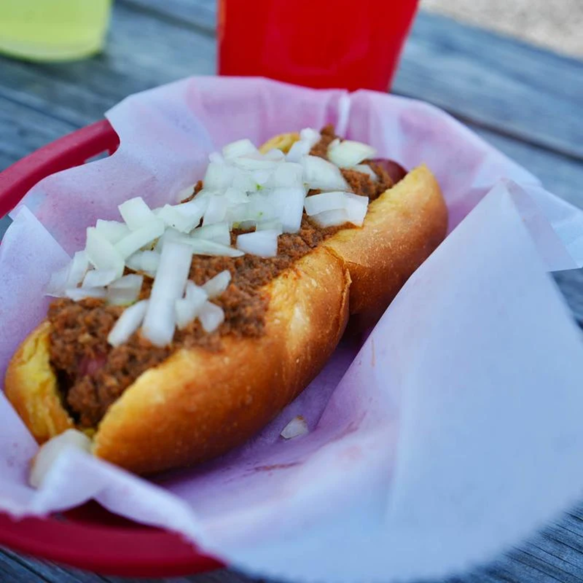A chili dog topped with chopped onions sits in a red basket lined with white paper on a wooden table, with colorful drinks blurred in the background.