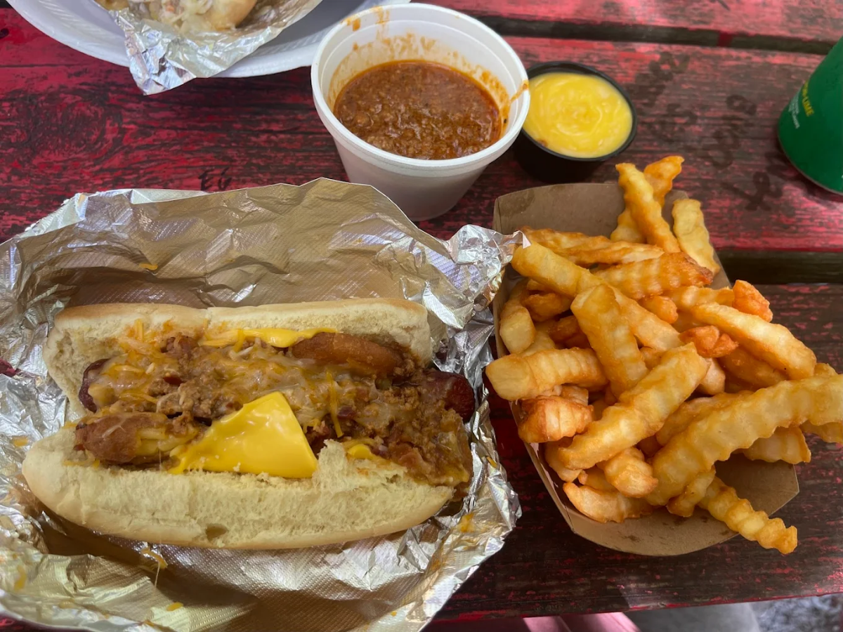 A hot dog topped with chili and cheese in a bun, served with crinkle-cut fries in a paper tray, a cup of chili, and a small container of yellow cheese sauce on a red wooden table.