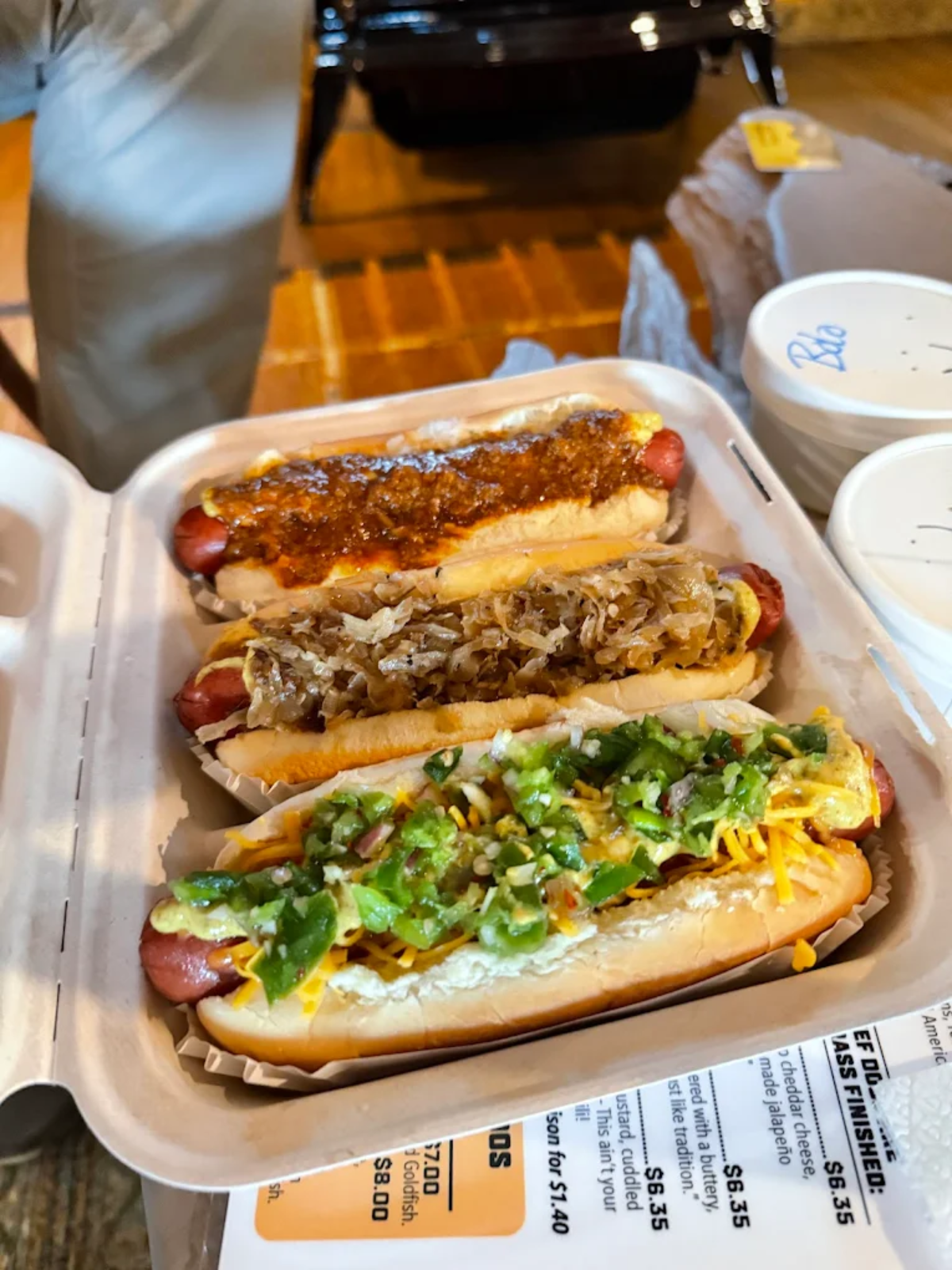 Three hot dogs in a takeout container, each topped with different ingredients: chili, sautéed onions, and a mix of cheese, jalapeños, and diced onions. A receipt and two lidded cups are also visible.