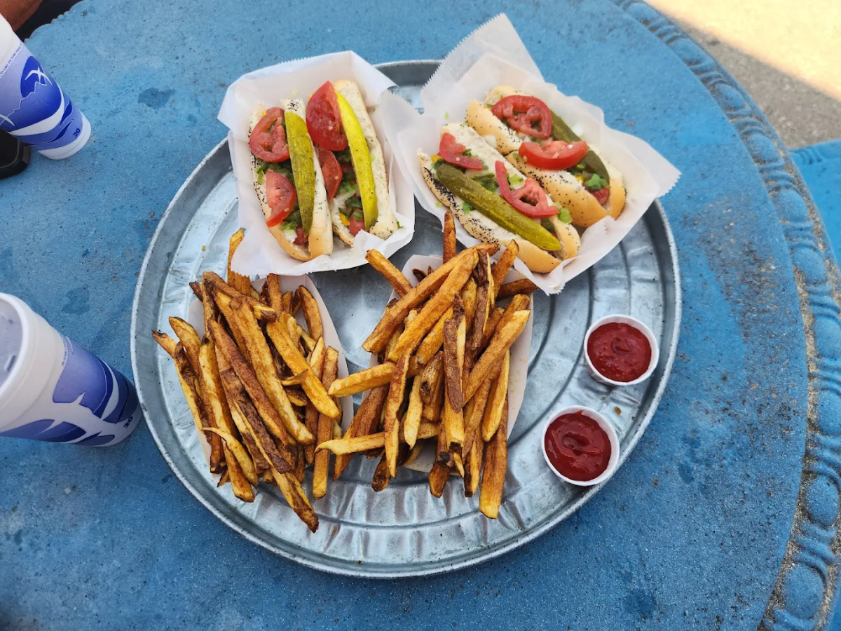 Two Chicago-style hot dogs with tomato slices, pickles, and relish sit on a metal tray with a large serving of fries, two cups of ketchup, and two blue-and-white soft drink cups on a blue table.