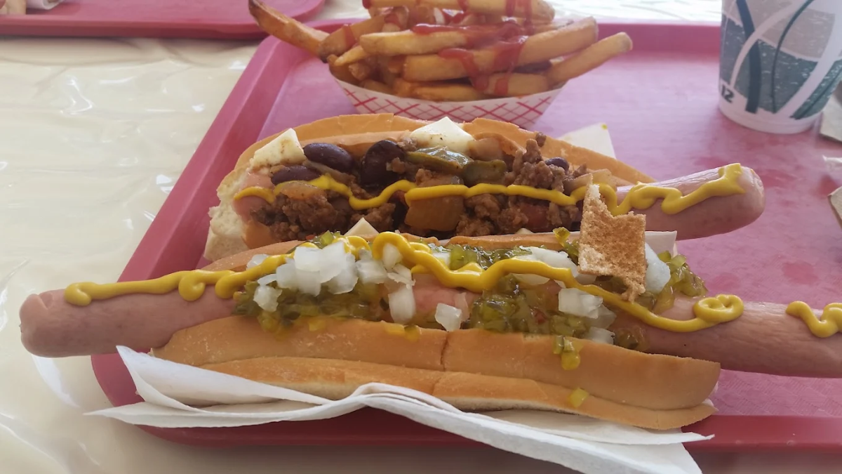 Two hot dogs on buns, one topped with chili, beans, onions, and chips, the other with mustard, relish, and onions. A serving of fries with ketchup and a drink cup are in the background on a red tray.