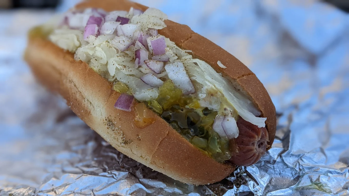 A hot dog in a bun topped with chopped onions, relish, and shredded sauerkraut, resting on crumpled aluminum foil.