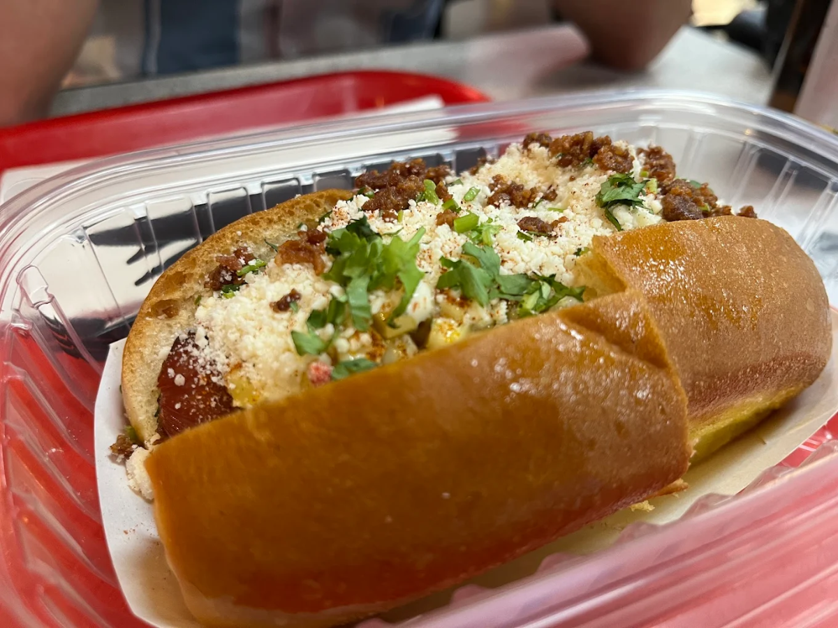 A close-up of a hot dog in a soft bun, topped with crumbled cheese, chopped cilantro, and crispy bits, served in a clear plastic container on a red tray.