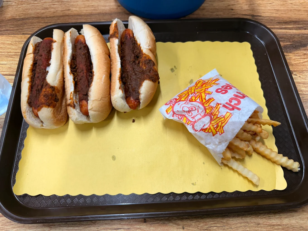 A tray with three chili dogs in buns and a small paper bag of crinkle-cut French fries on a yellow paper liner. The tray is resting on a wooden table.