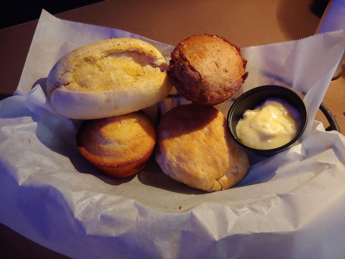 A basket lined with parchment paper holds a variety of bread and muffins, accompanied by a small black container of butter or spread.