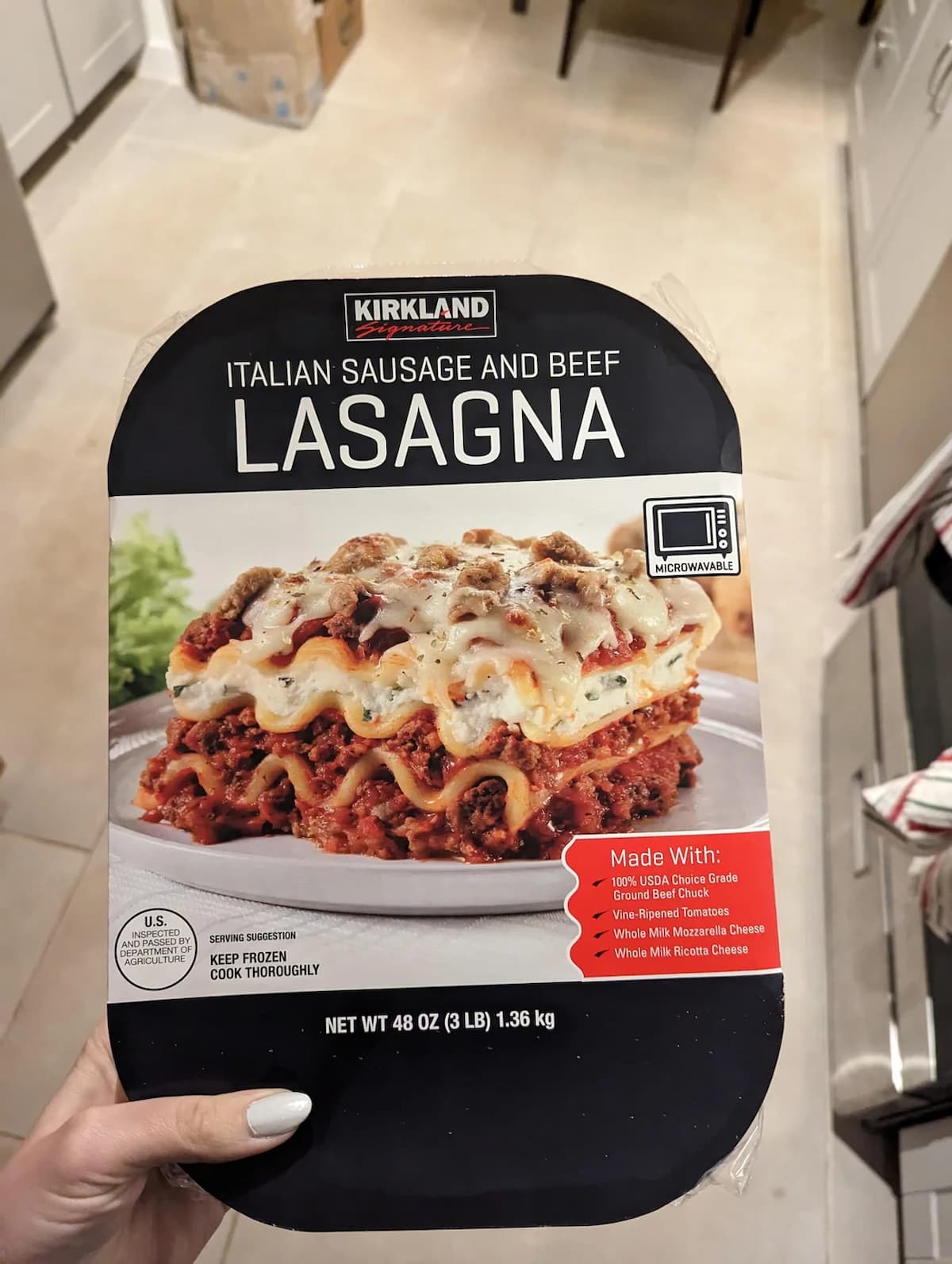A hand holds a package of Kirkland Italian Sausage and Beef Lasagna. The label shows a photo of a cooked lasagna slice and lists ingredients like USDA ground chuck, Italian sausage, mozzarella, and ricotta cheese.