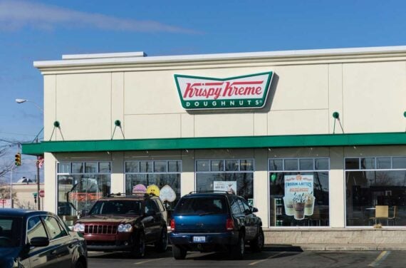 A Krispy Kreme Doughnuts shop with cars parked in front on a sunny day. The storefront features large windows, a green-and-red Krispy Kreme sign above the entrance, and a banner advertising special tax day deals.