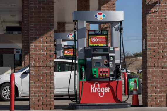 A white car is parked at a Kroger gas station pump under a brick canopy. The pump advertises cheap gas and promotions. Several pumps and red bollards are visible, with a clear blue sky in the background.