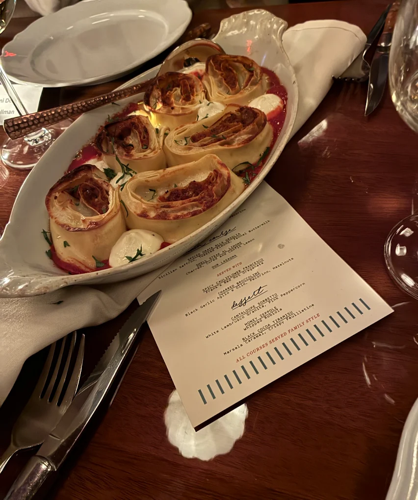 A close-up of a ceramic dish with rolled pasta in tomato sauce, garnished with herbs and cheese, placed on a wooden table alongside a restaurant menu, silverware, napkin, and wine glasses.
