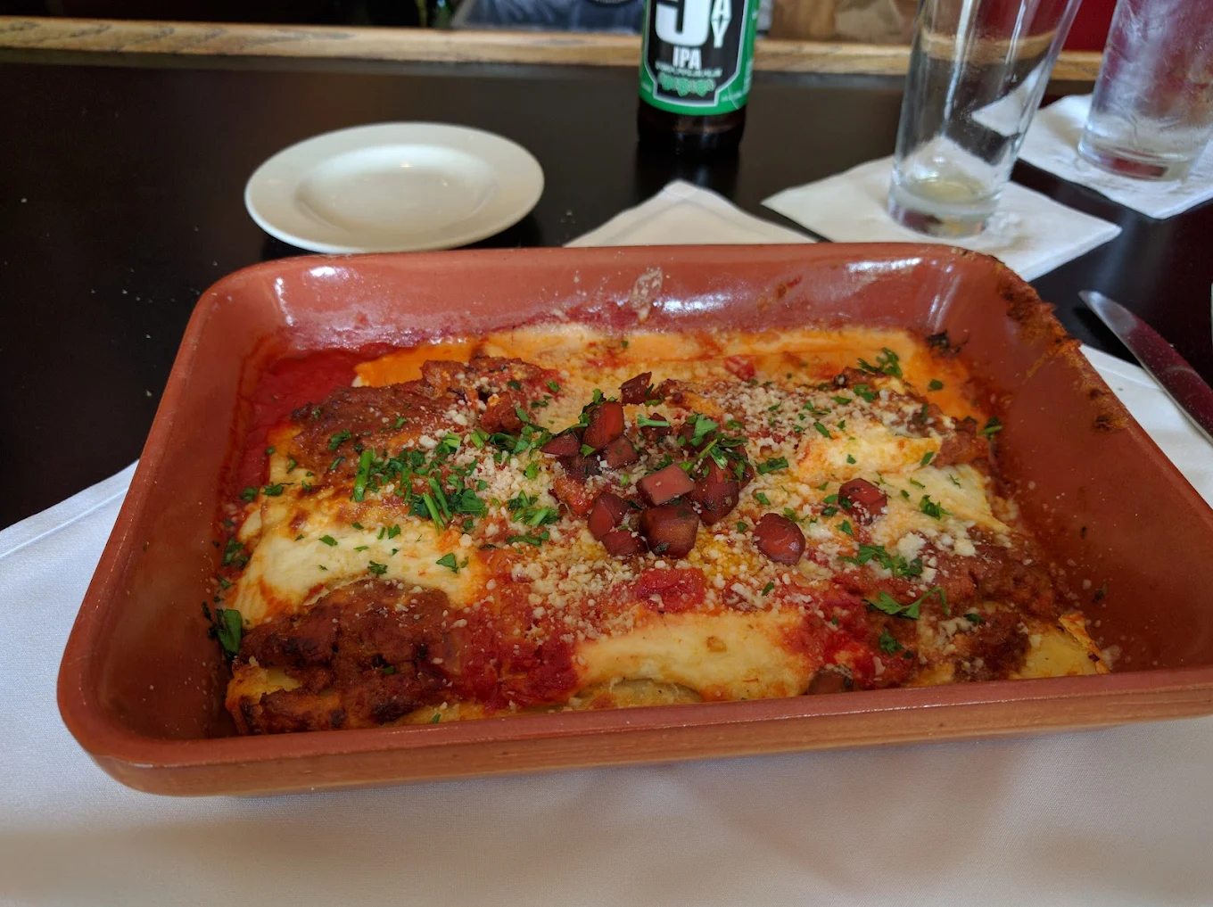 A baked dish of lasagna topped with tomato sauce, melted cheese, chopped herbs, and sliced olives, served in a rectangular ceramic dish on a table with an empty plate, glasses, and a bottled drink nearby.