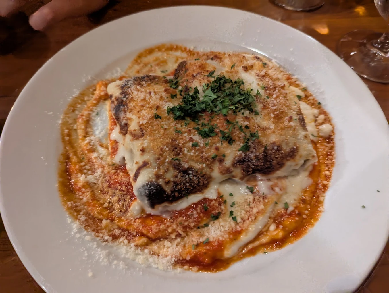 A plate of baked lasagna topped with melted cheese, tomato sauce, breadcrumbs, grated parmesan, and chopped parsley, served on a white dish.