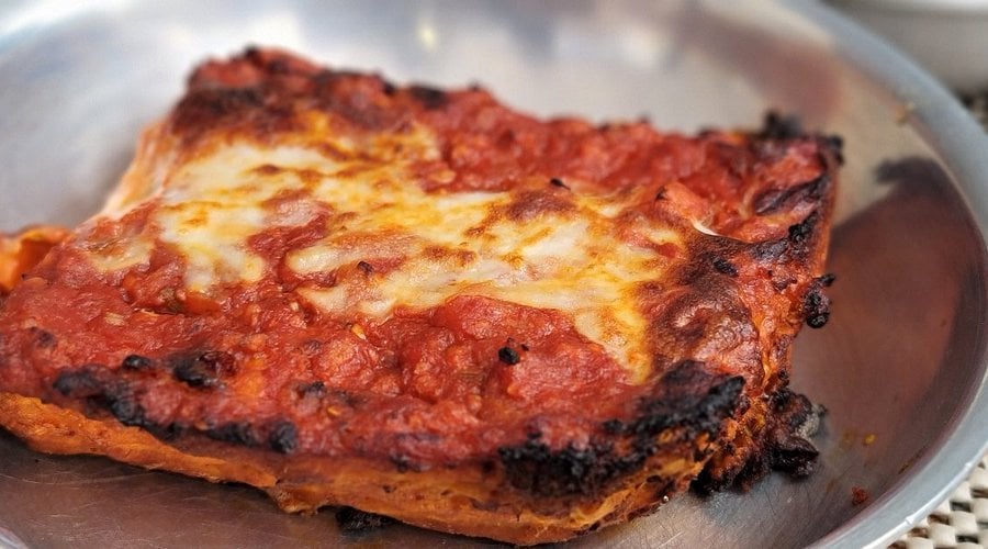 A close-up of a rectangular slice of lasagna with melted cheese and tomato sauce on top, served on a round metal plate. The edges are slightly browned and crisp.
