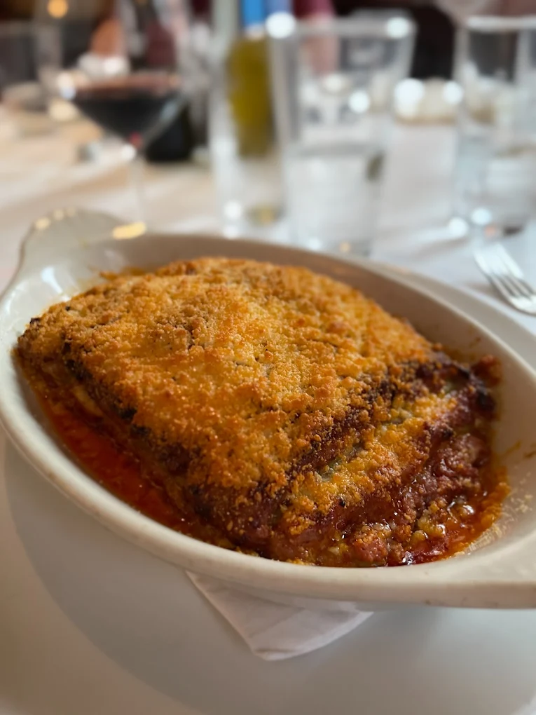 A close-up of a baked dish, likely lasagna or eggplant parmesan, in a white oval dish with a golden, crispy breadcrumb topping, on a white tablecloth with glasses and cutlery in the background.