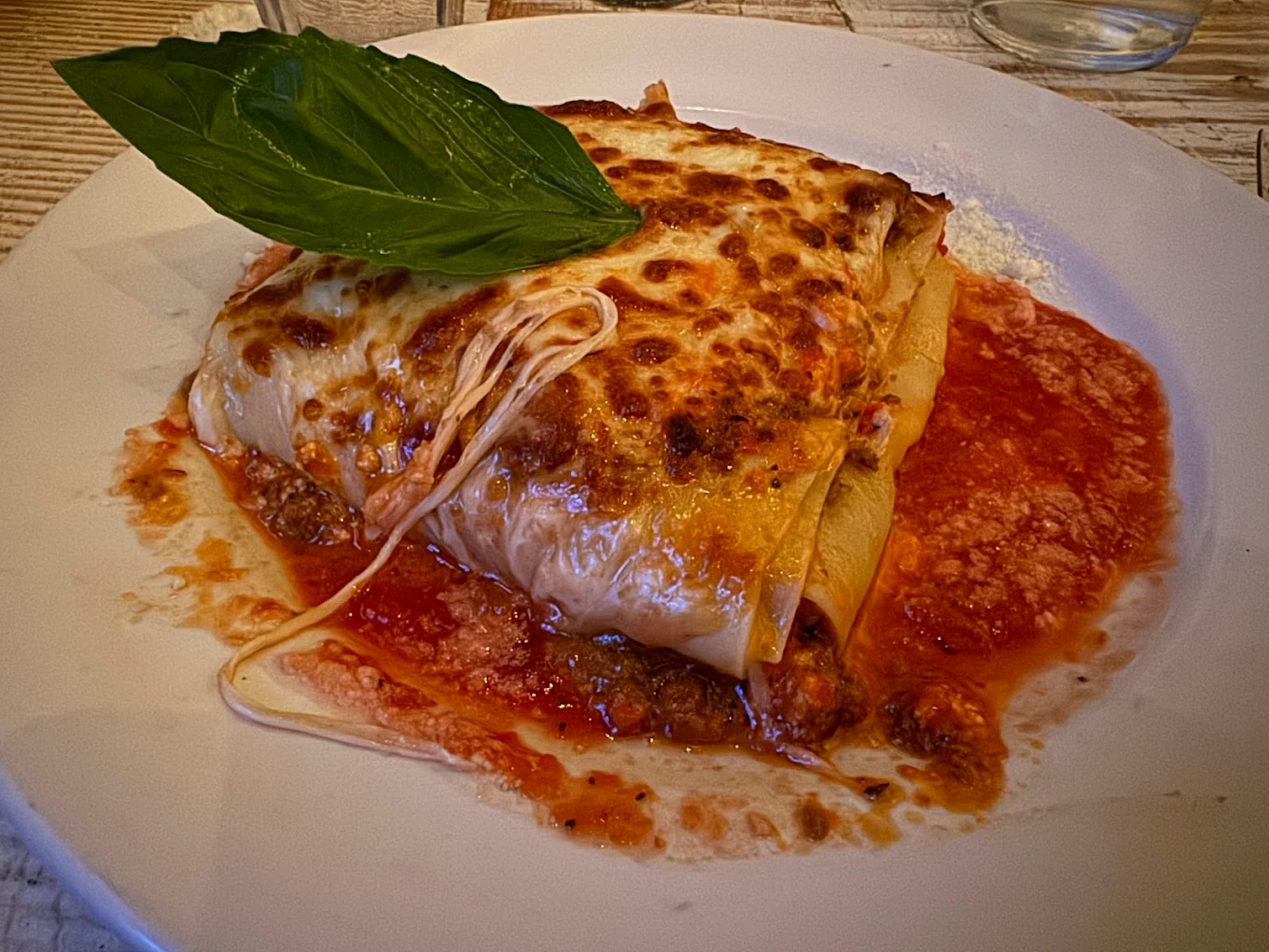 A serving of cheesy baked lasagna sits on a white plate, topped with a fresh basil leaf and surrounded by tomato sauce. Melted cheese is slightly browned, and the lasagna layers are visible with meat and sauce.