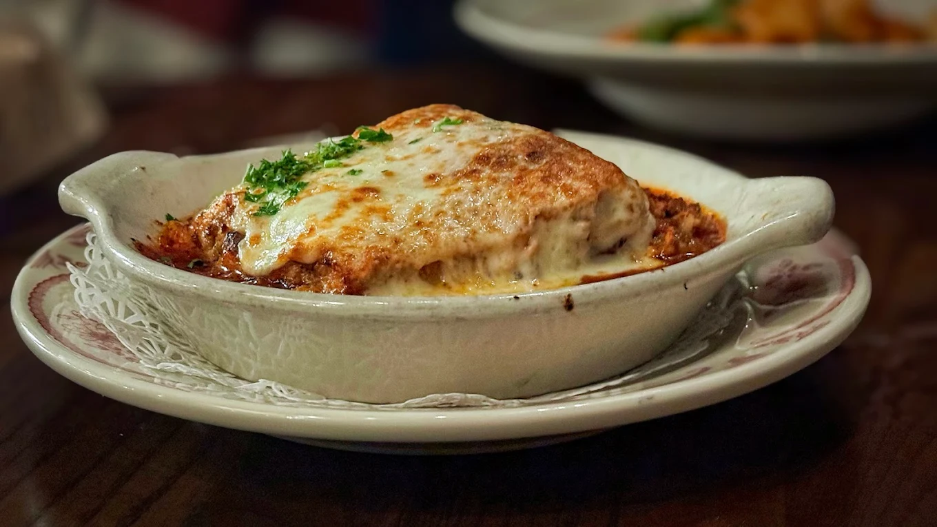 A close-up of a baked lasagna in a white ceramic dish, topped with melted, golden-brown cheese and garnished with parsley, sitting on a decorative plate on a wooden table.