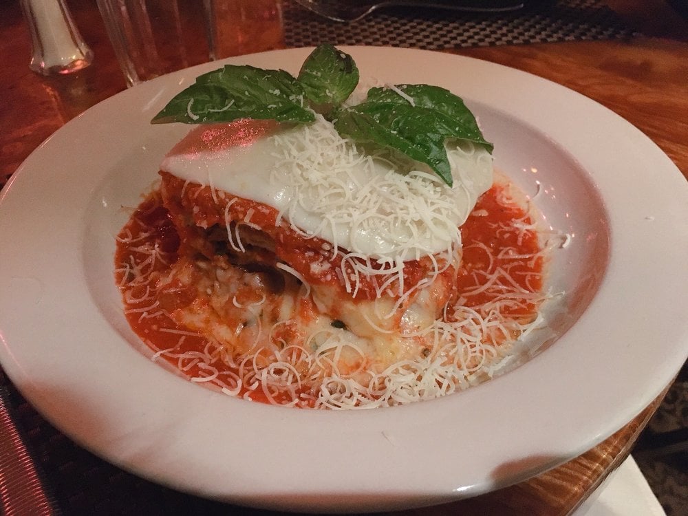 A serving of lasagna topped with melted cheese, shredded Parmesan, and fresh basil leaves, sits on a white plate surrounded by marinara sauce.