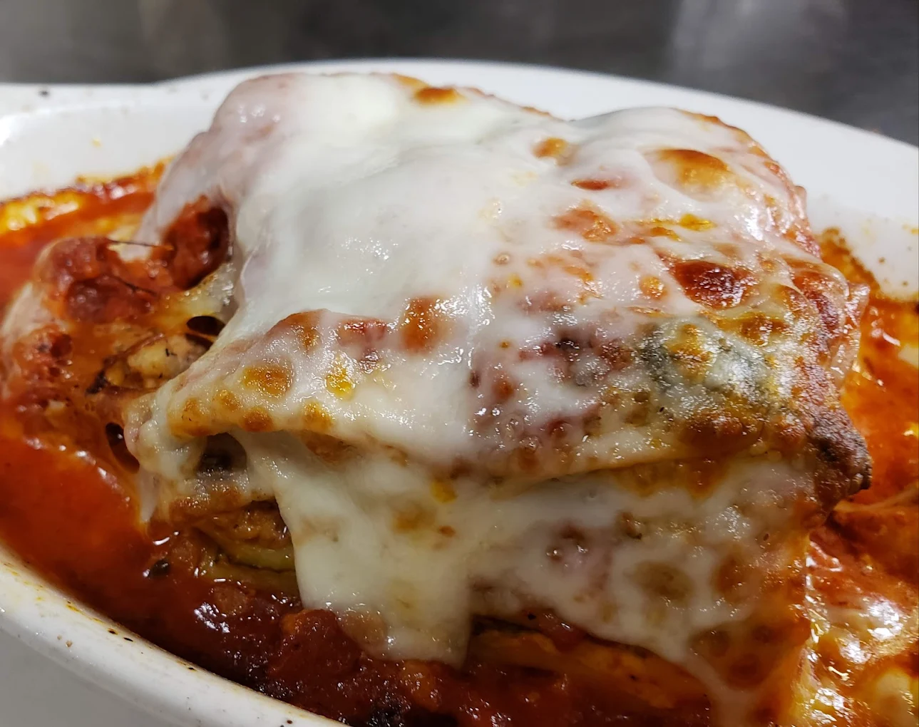 A close-up of a serving of lasagna topped with melted cheese in a white dish, with layers of pasta, meat, sauce, and cheese visible, sitting in a pool of rich red marinara sauce.