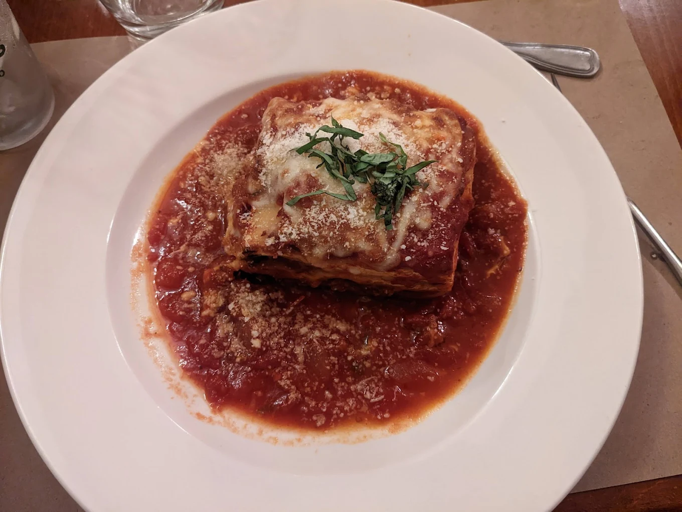 A slice of lasagna topped with grated cheese and fresh basil sits on a white plate, surrounded by a generous serving of tomato sauce. A glass and cutlery are partially visible in the background.