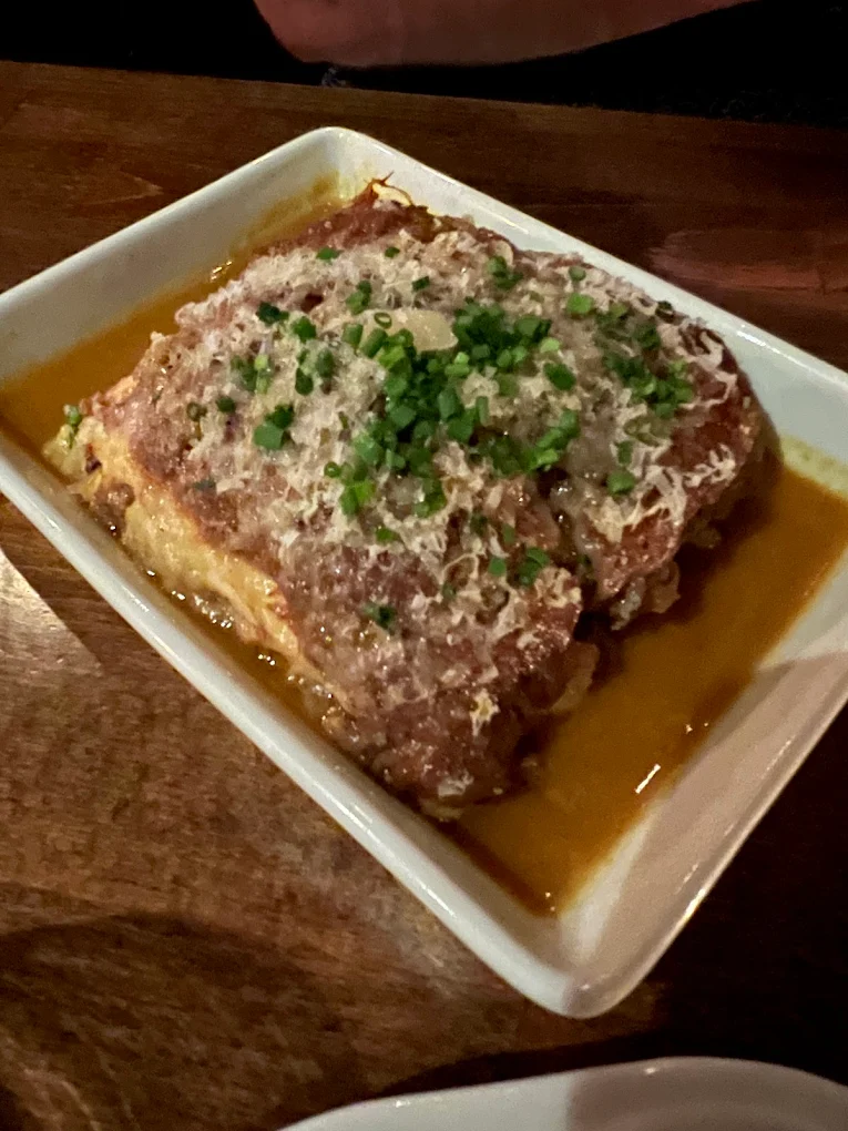A rectangular piece of lasagna topped with grated cheese and chopped chives sits in a white dish with a pool of sauce, on a wooden table.